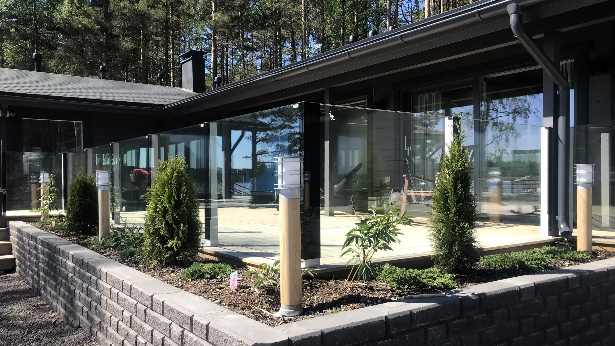 Moderni kiinteä lasikaide omakotitalon pihassa – lasielementti istutusten ja kivetyksen yhteydessä.